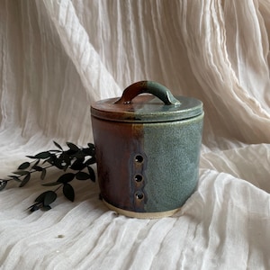 May include: A handmade ceramic jar with a lid and handle. The jar is a mix of brown and green colours, with three small holes on the side. The lid has a curved handle. The jar is likely used for storage.