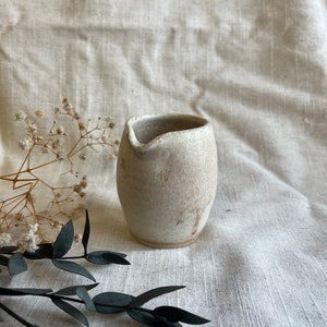 May include: A small, white ceramic jug with a speckled glaze. The jug has a rounded body and a slightly flared lip.