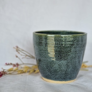 Könnte beinhalten: Eine dunkelgrüne Keramiktasse mit einem gesprenkelten Überzug. Die Tasse hat eine abgerundete Form und eine leicht ausgestellte Lippe.