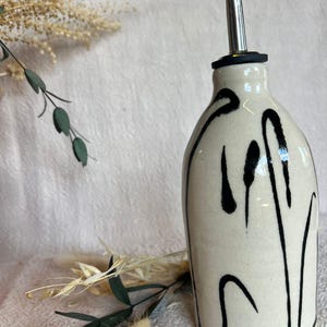 K&ouml;nnte beinhalten: Cremefarbene &Ouml;lflasche aus Keramik mit Edelstahlausguss und schwarzem, abstraktem Design. Die Flasche hat eine abgerundete Form und ist mit vertikalen und geschwungenen schwarzen Linien verziert. Das Design verleiht der Flasche ein modernes, k&uuml;nstlerisches Aussehen.