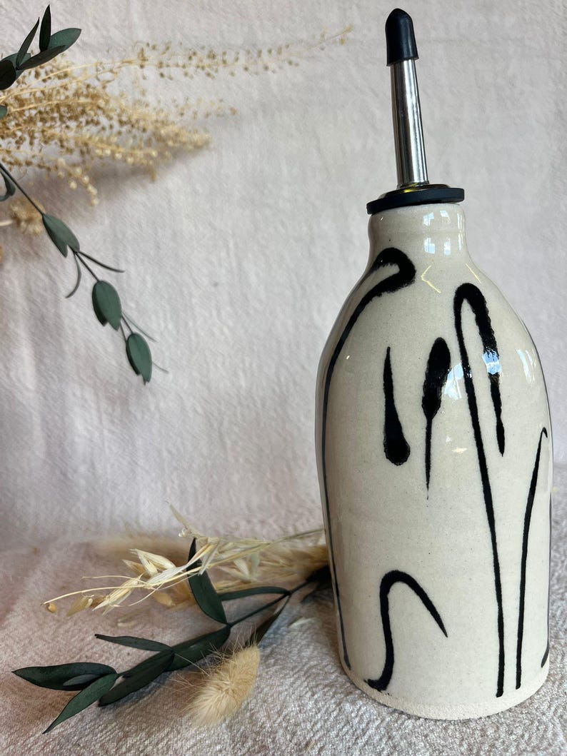 K&ouml;nnte beinhalten: Cremefarbene &Ouml;lflasche aus Keramik mit Edelstahlausguss und schwarzem, abstraktem Design. Die Flasche hat eine abgerundete Form und ist mit vertikalen und geschwungenen schwarzen Linien verziert. Das Design verleiht der Flasche ein modernes, k&uuml;nstlerisches Aussehen.