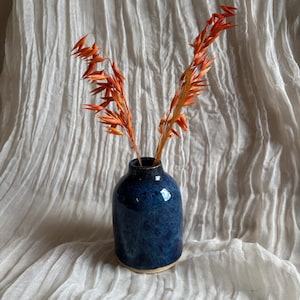 May include: A small, dark blue ceramic vase with a speckled glaze, holding two stems of orange dried flowers. The vase has a rounded shape and a light tan base, set against a white, textured fabric background.
