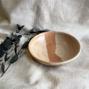 May include: A small, round ceramic bowl with a white, yellow, and brown glaze. The bowl is sitting on a light brown fabric surface.