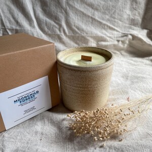 May include: A beige ceramic candle with a wooden wick, in a cylindrical shape. The candle is next to a brown box with the text "MOONCHILD FOREST". A small bundle of dried flowers is next to the candle.
