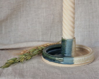 May include: A handmade ceramic candle holder with a cream-colored twisted candle. The holder is divided into sections of green, blue, and beige. A sprig of wheat is placed next to the holder, adding a rustic touch.