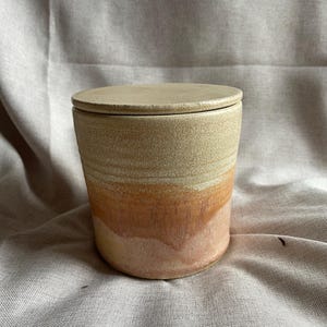 May include: A ceramic jar with a lid, featuring a light beige glaze with a band of orange and pink glaze around the middle.