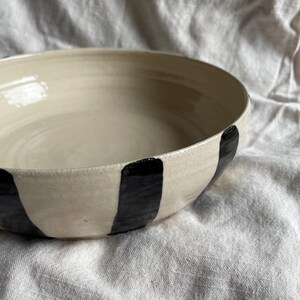May include: A large, white ceramic bowl with black vertical stripes. The bowl is handmade and has a rustic, organic feel.