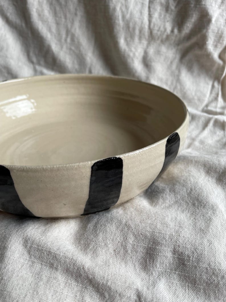 May include: A large, white ceramic bowl with black vertical stripes. The bowl is handmade and has a rustic, organic feel.