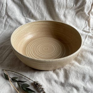 May include: A large, beige ceramic bowl with a textured, swirling pattern. The bowl is sitting on a white fabric surface.