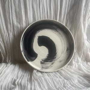 May include: A circular ceramic plate featuring a cream-coloured base and a striking black brushstroke design. The black paint swirls across the plate's surface, creating a bold contrast. The plate is set against a white, textured background.