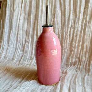 May include: A glossy pink ceramic oil cruet with a smooth, rounded body and a narrow neck. The top features a silver-colored metal pour spout with a black rubber seal, designed for dispensing liquids from the bottle's interior container.