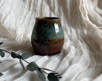 May include: A small, handcrafted ceramic vase with a glossy finish. The vase features a gradient of colors, transitioning from a dark brown base to a teal-green midsection and a dark brown rim. The vase is set against a soft, white fabric background.