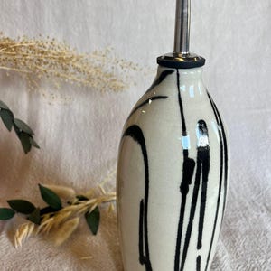K&ouml;nnte beinhalten: Eine Keramik&ouml;lflasche mit einem schwarz-wei&szlig;en Design. Die Flasche hat einen Ausgie&szlig;er aus Edelstahl mit einer schwarzen Gummit&uuml;lle. Die Flasche ist hellbeige mit schwarzen Farbtropfen.