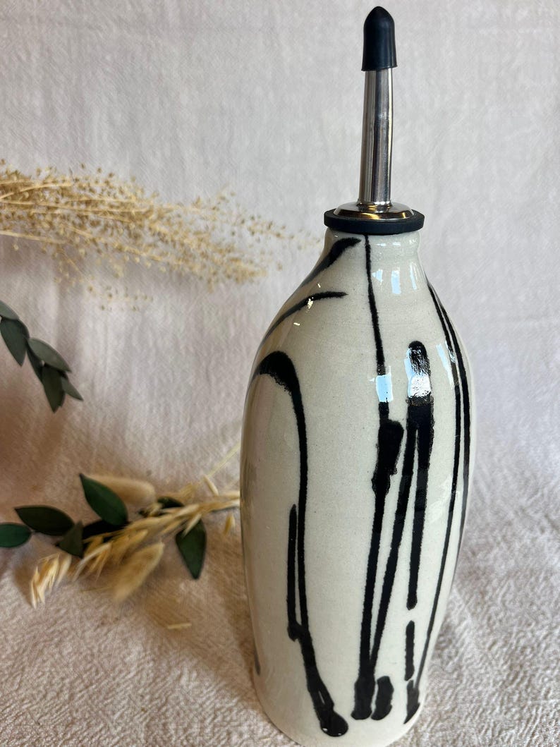 K&ouml;nnte beinhalten: Eine Keramik&ouml;lflasche mit einem schwarz-wei&szlig;en Design. Die Flasche hat einen Ausgie&szlig;er aus Edelstahl mit einer schwarzen Gummit&uuml;lle. Die Flasche ist hellbeige mit schwarzen Farbtropfen.