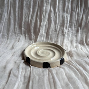 May include: A cream-coloured ceramic soap dish with black polka dots around the rim. The dish features a spiral design in the centre. It is set against a white, textured fabric background.