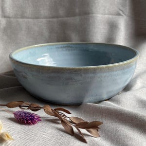May include: A light blue ceramic bowl with a slightly darker rim. The bowl is sitting on a light grey fabric surface.