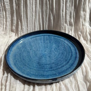 May include: A round, ceramic plate with a glossy, textured blue surface and a dark blue rim. The plate has a slightly raised edge and is set against a white, textured background. The plate is likely for serving food.