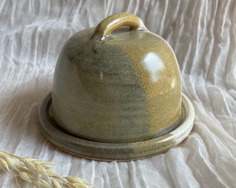 May include: A handmade ceramic butter dish with a lid and handle. The dish is a dome shape, with a base and lid in shades of green and tan. The handle is arched. The dish is sitting on a white, textured surface.