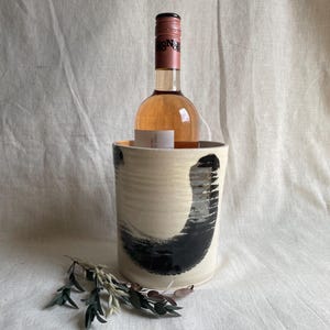 May include: A white ceramic wine cooler with black abstract brushstrokes. The cooler is holding a bottle of rose wine.