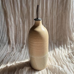 May include: A beige ceramic oil bottle with a stainless steel spout. The bottle has a textured, speckled finish on the upper portion and a smooth, cream-coloured base. The design is simple and elegant, suitable for kitchen use.