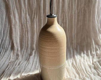 May include: A beige ceramic oil bottle with a stainless steel spout. The bottle has a textured, speckled finish on the upper portion and a smooth, cream-colored base. The design is simple and elegant, suitable for kitchen use.