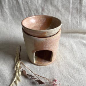 May include: A ceramic essential oil diffuser with a light pink and white glaze. The diffuser has a small opening on the side for a tea light candle.
