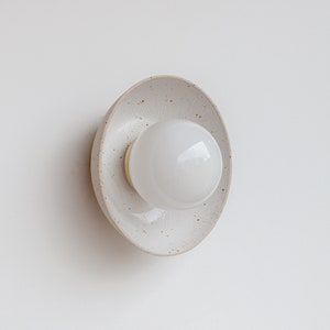 Può includere: Un'applique da parete in ceramica bianca con finitura macchiettata e una lampadina rotonda bianca.
