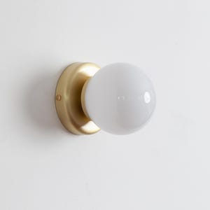 Könnte beinhalten: Eine goldene Wandleuchte mit einer weißen Kugellampe. Die Leuchte hat ein schlichtes, modernes Design.