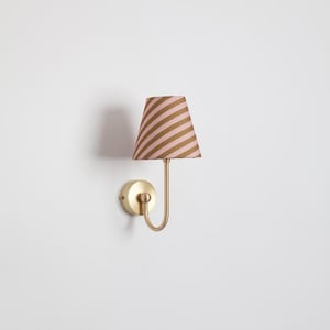 Puede incluir: Aplique de pared con acabado dorado. La pantalla es de forma cónica con rayas diagonales rosas y doradas. El aplique tiene un brazo curvo y una base circular.