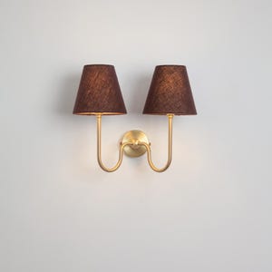 Puede incluir: Aplique de pared doble con dos pantallas de tela marrón oscuro. La lámpara tiene una base de metal dorado y brazos curvos. Las pantallas son de color marrón oscuro y la lámpara está montada en una pared blanca.