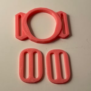 Pode incluir: Três fivelas deslizantes de plástico rosa. A fivela maior tem uma abertura redonda e duas abas de cada lado. As duas fivelas menores são retangulares com duas ranhuras.