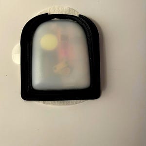 May include: A close-up of a white, round, adhesive patch with a black border. The patch has a clear, plastic window that reveals a small, yellow object and other components.