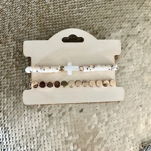 May include: Two bracelets displayed on a light wood card. The top bracelet features white beads with black spots and a white cross. The bottom bracelet has gold-colored disc beads. The card has a hole at the top for hanging.