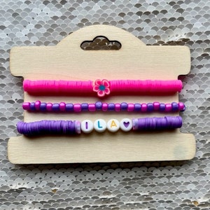 May include: Three bracelets on a wooden display card. The top bracelet is pink and has a flower charm. The middle bracelet is purple and has small beads. The bottom bracelet is purple and has the name "ILA" spelled out with colorful beads and a heart charm.