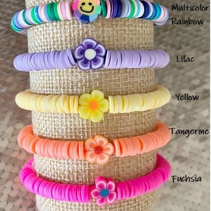 May include: Five colorful bracelets with flower charms. The bracelets are stacked on a burlap background. The colors of the bracelets are rainbow, lilac, yellow, tangerine, and fuchsia. The text "Multicolor Rainbow", "Lilac", "Yellow", "Tangerine", and "Fuchsia" are written next to each bracelet.