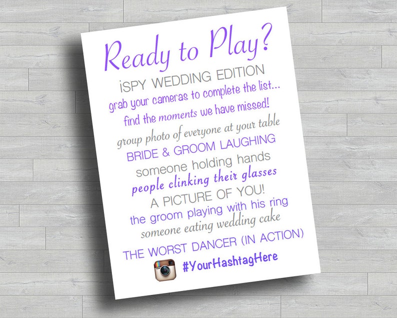 May include: A printable wedding game card with purple text that says "Ready to Play? iSpy Wedding Edition" and lists a series of wedding-themed photo challenges for guests to complete. The card also includes a hashtag for the wedding.