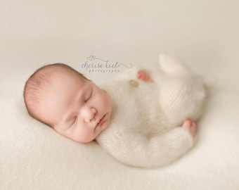 May include: A newborn baby is sleeping soundly in a white knitted onesie. The baby is lying on a white fluffy blanket. The baby's face is visible and the eyes are closed.