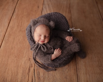 May include: A newborn baby wearing a gray knitted hat and matching outfit is sleeping in a gray knitted basket. The baby is lying on its back with its arms crossed over its chest. The basket is sitting on a wooden floor.