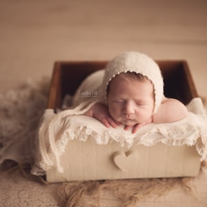 May include: A newborn baby wearing a white crocheted hat is sleeping in a white wooden box with a heart cutout. The box is lined with a white lace fabric.