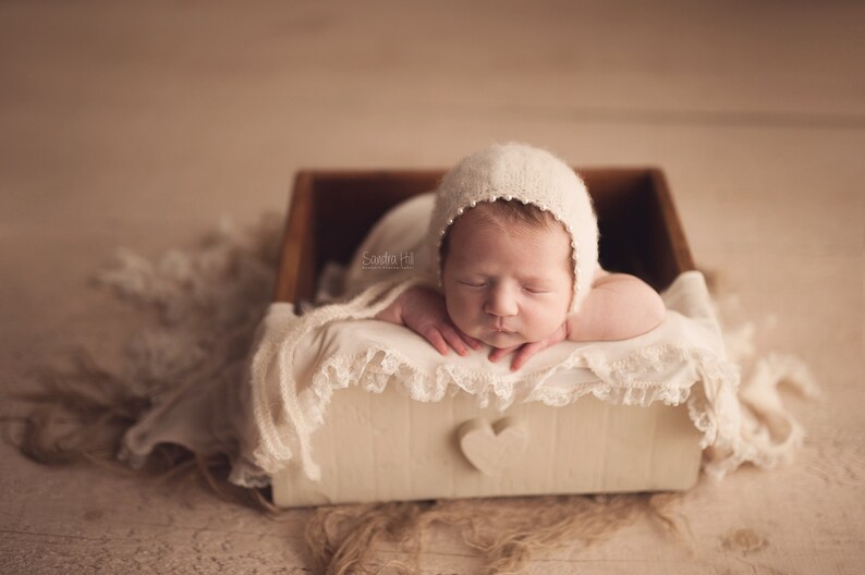 May include: A newborn baby wearing a white crocheted hat is sleeping in a white wooden box with a heart cutout. The box is lined with a white lace fabric.