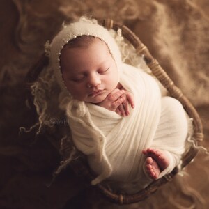 May include: A newborn baby is swaddled in a white blanket and wearing a white crocheted bonnet with pearl accents. The baby is sleeping peacefully in a wicker basket.