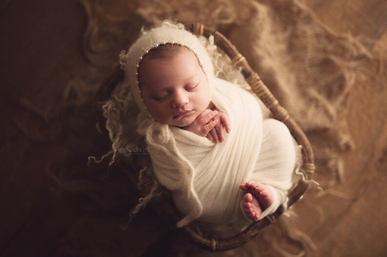 May include: A newborn baby is swaddled in a white blanket and wearing a white crocheted bonnet with pearl accents. The baby is sleeping peacefully in a wicker basket.