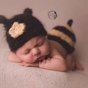 Op de afbeelding: Een pasgeboren baby in een zwart en geel bijen-thema outfit, inclusief een hoed met oren en een bloemaccent. De baby ligt op een getextureerd oppervlak, met gesloten ogen. De outfit is handgemaakt en geschikt voor een fotoshoot.