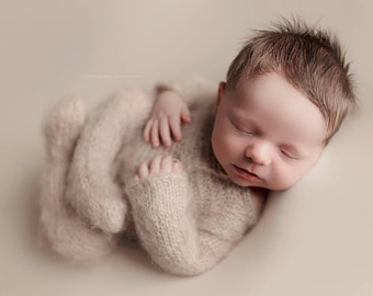 May include: A newborn baby is sleeping soundly in a soft, fuzzy, light brown knitted onesie. The baby has a full head of brown hair and is curled up in a peaceful slumber.