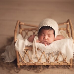 May include: A newborn baby wearing a white crocheted bonnet is sleeping in a small wicker crib. The crib is lined with a white fabric and the baby is resting on its arms.