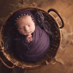 May include: A newborn baby is sleeping in a woven basket. The baby is wrapped in a purple swaddle and wearing a white hat with pearl accents. The basket is made of natural materials and has a brown color.