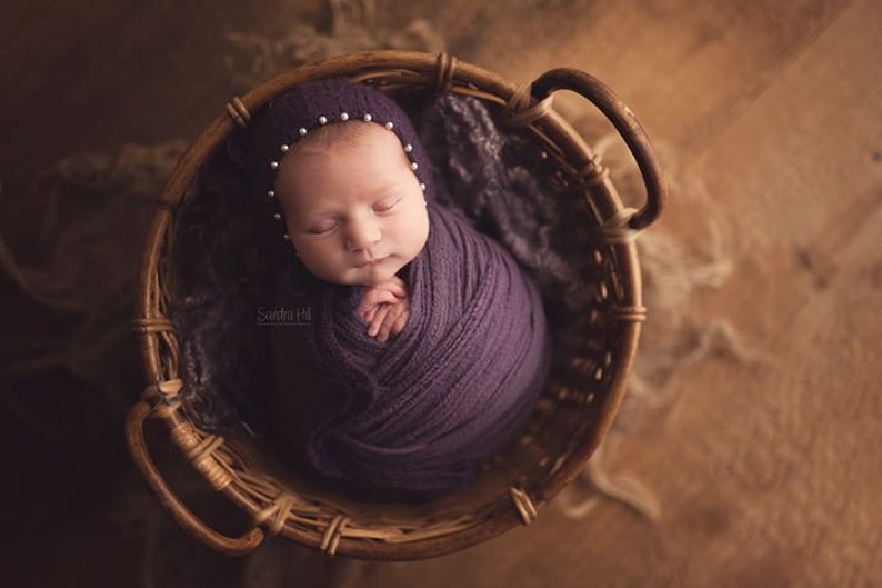 May include: A newborn baby is sleeping in a woven basket. The baby is wrapped in a purple swaddle and wearing a white hat with pearl accents. The basket is made of natural materials and has a brown color.