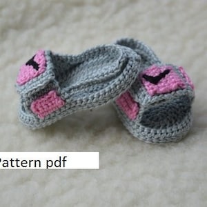 May include: A pair of gray crocheted baby sandals with pink and black accents. The sandals have a strap across the top and a small pink heart on the side. The text "Pattern pdf" is visible in the image.
