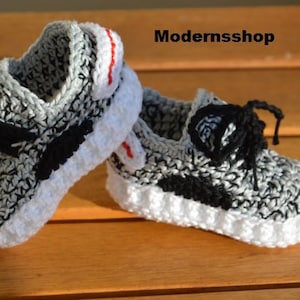 May include: A pair of gray, black, and white crocheted baby shoes with black laces. The shoes have a white sole and a red accent on the back.