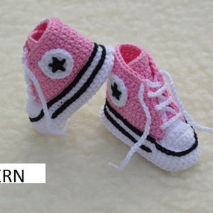 Puede incluir: Un par de zapatos de bebé de crochet rosa con ribetes blancos y negros, que se asemejan a las zapatillas Converse. Los zapatos tienen un diseño de estrella en el lateral.